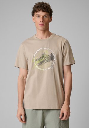 Młody mężczyzna w beżowym t-shircie z grafiką mapy gór i napisem „Im wyżej, tym lepiej”, stojący na jednolitym szarym tle.