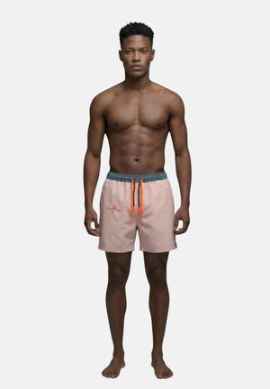Homme athlétique debout pieds nus, portant un short de bain rose clair avec une ceinture foncée et des cordons orange sur un fond uni.