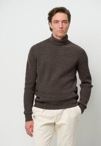 Brauner gerippter Rollkragenpullover aus einem weichen Wollgemisch, taillierter Schnitt, mit umgeschlagenem Kragen und langen Ärmeln, kombiniert mit hellen Hosen.