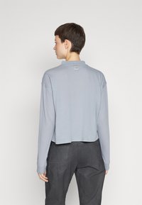 Cămașă scurtă cu mâneci lungi în gri deschis, având un guler înalt și o etichetă mică pe spate. Material moale cu o textură fină.
