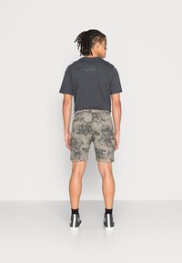 Camiseta preta de manga curta com o logotipo "adidas", combinada com calções cinzentos de camuflagem. Ténis altos pretos com detalhes brancos. Fundo simples.