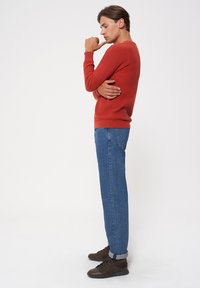 Czerwony sweter o fakturze, dopasowany krój, okrągły dekolt, sparowany z niebieskimi dżinsami typu straight-leg i brązowymi butami, stojący w profilu bocznym na tle jednolitego tła.