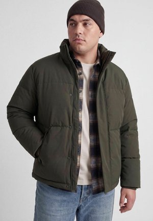 Homme portant une doudoune vert foncé, une chemise à carreaux, un t-shirt blanc, un jean bleu et un bonnet en tricot marron, debout avec une main dans la poche de la veste.