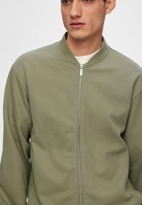 Veste zippée vert olive avec col montant, en tissu lisse et à texture subtile, conçue pour un look moderne et minimaliste.