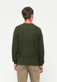 Uomo di spalle che indossa un maglione a maglia verde scuro e pantaloni marrone chiaro su uno sfondo bianco semplice.