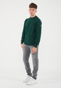 Maglione verde lavorato a maglia con maniche lunghe, scollo rotondo e un piccolo patch sulla manica sinistra, abbinato a jeans grigi e sneakers bianche.