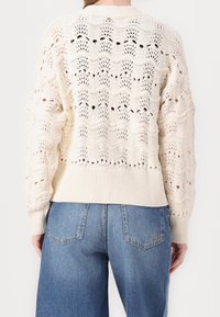 Vit stickad tröja med ett mönster i öppen väv och ribbade muddar, med en vågmönsterdesign och texturdetalj på ryggen. Kombineras med blå jeans.