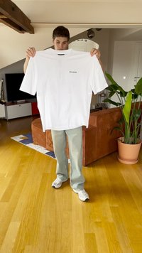 Biela tričko s krátkymi rukávmi a potlačeným logom. Model ho drží pred pozadím s drevenou podlahou a izbovými rastlinami.