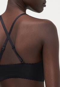 Black bra, featuring crisscross straps, a smooth texture, and scalloped edges. Minimal hardware accents near strap adjusters.