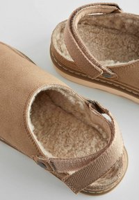 Tan fabric shoes with a soft, textured interior, open design, and adjustable strap feature faded stitching and a flat rubber sole.