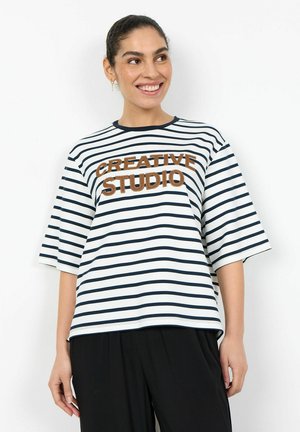 Donna sorridente con capelli scuri raccolti in una coda di cavallo, che indossa una maglietta a righe bianche e blu con la scritta "CREATIVE STUDIO" e pantaloni neri.