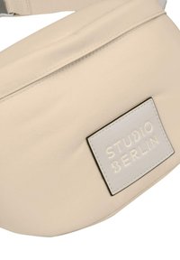 Marsupio beige realizzato in materiale morbido, caratterizzato da un grande logo in rilievo argentato e una tracolla regolabile con dettagli di cucitura.