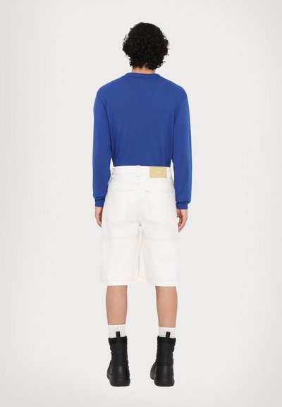 Top de manga larga azul, pantalones cortos de mezclilla blancos con costuras visibles y botas negras hasta el tobillo. La etiqueta en la cintura es de color claro con texto.