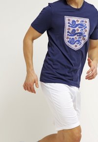 Marineblaues T-Shirt mit kurzen Ärmeln, das ein großes Wappenmotiv mit Löwen in Blau und Rot zeigt, kombiniert mit weißen Sportshorts.