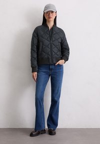 Czarna pikowana kurtka bomberka z prążkowanym mankietem, zestawiona z niebieskimi jeansami z rozszerzanymi nogawkami oraz ciemnymi mokasynami. Szara czapka dopełnia strój.