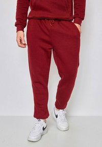 Pantalons de survêtement rouges en tissu doux, avec une taille élastique et un cordon de serrage, et des jambes fuselées. Portés avec des baskets blanches.