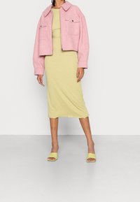 Valkoinen crop-mallinen takki, jossa on kaksi etutaskua, keltaisen raidallisen midi-mekon päällä. Vaaleanvihreät blokkikorkoiset liukulevyn sandaalit viimeistelevät asun.