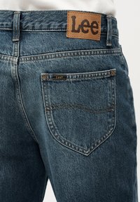 Πίσω όψη ατόμου που φοράει μπλε τζιν παντελόνι της μάρκας Lee, με δερμάτινη ετικέτα Lee στη μέση και μικρή ετικέτα Lee στην πίσω τσέπη.