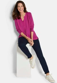 Top magenta a maniche lunghe con scollo a V e leggera arricciatura sulle spalle, abbinato a pantaloni slim-fit blu scuro e sneakers bianche.