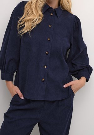 Femme portant un chemisier bleu marine à boutons avec des manches bouffantes et un pantalon assorti bleu marine, les mains dans les poches, sur un fond uni.