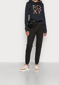 Persona vestida con una sudadera con capucha negra con el texto "ROXY" estampado, pantalones de chándal negros, zapatillas rosa y beige, y una riñonera negra.