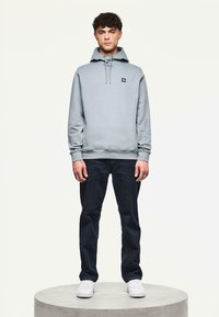 Felpa grigia con cappuccio e un piccolo logo nero, abbinata a jeans scuri e sneakers bianche. Il modello si trova su un piedistallo circolare.