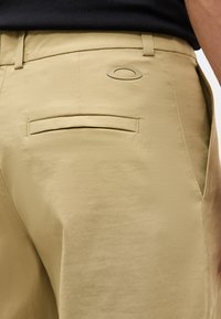 Κοντινή λήψη πίσω τσέπης μπεζ παντελονιού με θηλιές ζώνης, μια οριζόντια σχισμή τσέπης και ένα οβάλ λογότυπο πάνω από την τσέπη στη δεξιά πλευρά.