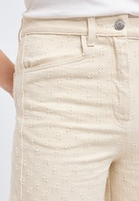 Κοντινή λήψη μπεζ παντελονιού με μικρή εξωτερική κεντητή λουλουδένια διακόσμηση, μπροστινή τσέπη, θηλιά για ζώνη και μεταλλικό κουμπί κλεισίματος.