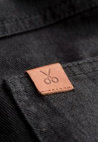 Tissu noir avec une étiquette en cuir marron présentant un design de ciseaux en relief. Les coutures sont visibles, mettant en avant la durabilité et le savoir-faire.