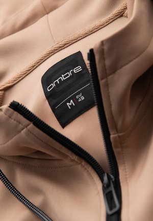Beige takki mustalla vetoketjulla osittain auki, näyttää mustan vaatemerkin "ombre" ja koon M eur 48.