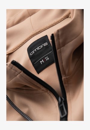 Beige takki mustalla vetoketjulla osittain auki, näyttää mustan vaatemerkin "ombre" ja koon M eur 48.