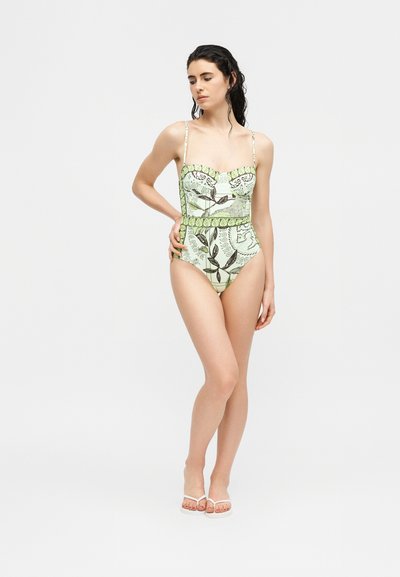 Femme debout en maillot de bain une-pièce vert à motifs floraux avec bretelles spaghetti et sandales blanches à talons, sur un fond uni.
