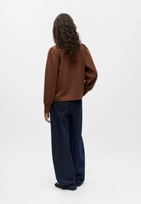Brauner, langärmliger Pullover mit puffärmligen und elastischen Bündchen, kombiniert mit hochgeschnittenen, weit geschnittenen dunkelblauen Jeans.