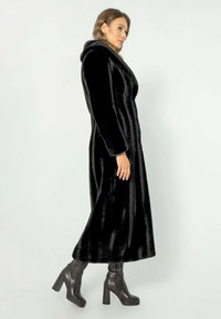 Černý kabát z umělé kožešiny s kapucí, který má elegantní siluetu a široké klopy, doplněný nadkolenními černými koženými botami na vysokém podpatku.