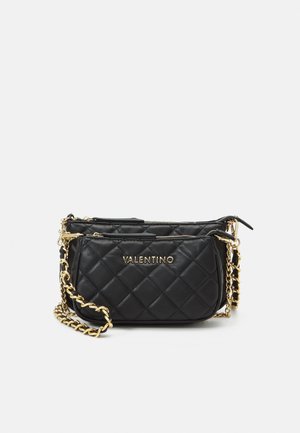 Zwarte doorgestikte Valentino crossbodytas met gouden kettingriem en gouden ritsdetails op een lichte achtergrond.