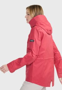Rote wasserdichte Jacke mit Kapuze, langen Ärmeln und Seitentaschen. "Aigle"-Logo am linken Arm. Glatte Stofftextur, taillierte Passform.