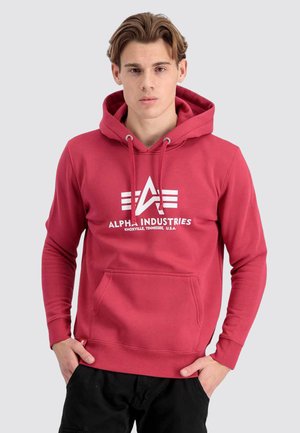 Junger Mann trägt einen roten Alpha Industries Hoodie mit Fronttasche, steht mit den Händen in den Taschen seiner schwarzen Hose vor einem einfarbigen Hintergrund.