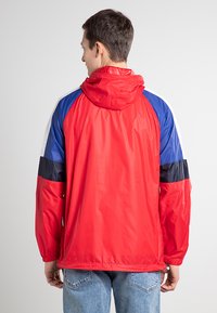 Veste coupe-vent rouge avec des accents de manches bleues et noires, dotée d'une capuche et d'un matériau lisse et léger. La vue arrière ne montre aucun détail supplémentaire.