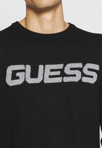 Schwarzer gestrickter Pullover mit einem auffälligen grauen "GUESS"-Logo auf der Vorderseite, klassischem Rundhalsausschnitt und gerippten Bündchen.
