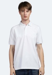 Polo shirt bianco con maniche corte, colletto abbottonato e tessuto texturizzato, indossato da un modello su uno sfondo chiaro.