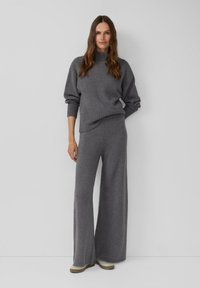 Woman with long brown hair wearing a matching grey knit sweater and wide-leg pants, standing against a plain light grey wall.