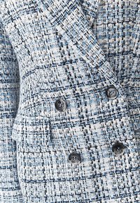 Blazer texturizado azul y blanco con un patrón de espiga, que presenta botones dobles y bolsillos laterales, hecho de tela tejida.