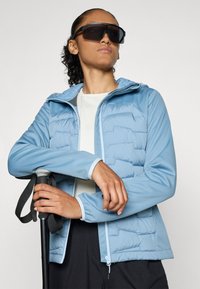 Lichtblauwe geïsoleerde jas met een getextureerde puffer voorkant, capuchon en aangesloten mouwen. Gecombineerd met een witte top en het vasthouden van een zwarte trekkingstok.
