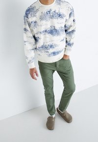 Mies, joka pitää päällä valkoista hupparia sinisellä maisemaprintillä, oliivinvihreitä housuja ja beigejä liukuesteitä, seisoo valkoista seinää vasten.