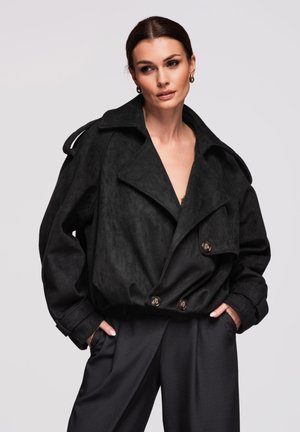 Kobieta w czarnej, oversize'owej, dwurzędowej marynarce z szerokimi klapami i czarnych spodniach, z rękami w kieszeniach, patrząca lekko na bok.