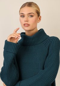 Maglione lavorato a maglia color teal con colletto alto e texture a coste. Maniche lunghe con polsini larghi, che mostrano cuciture dettagliate.