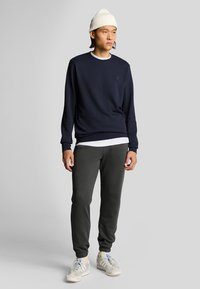 Felpa blu navy con polsini e colletto a costine, con un piccolo logo. Abbinata a pantaloni della tuta verde scuro e scarpe da ginnastica bianche. Berretto di maglia bianco.