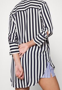 Personne portant une longue chemise rayée verticalement en noir et blanc avec les manches retroussées, tenant un poignet derrière le dos, montrant un aperçu d'un short rayé bleu.