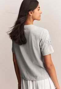 Lichtgrijs t-shirt met korte mouwen, gemaakt van een zachte stof en met een decoratieve geborduurde vlinder op de linker schouder.