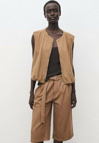 Beige ärmellose Weste mit einem Reißverschluss vorne, kombiniert mit passenden lockeren Shorts, die einen Gürtel und Seitentaschen haben. Strukturiertes Material.